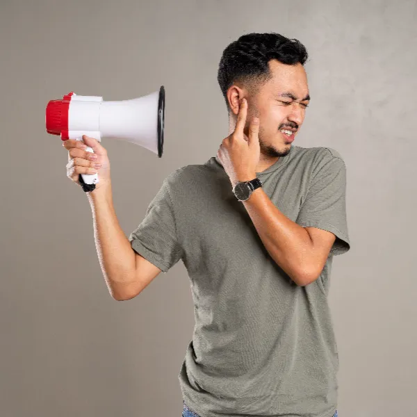 bundaberg-1 Man in a gray T-shirt shouting into a white megaphone with a red tip, covering one ear with his hand, against a neutral background.