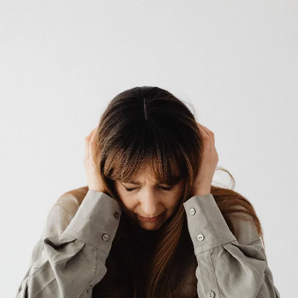 bundaberg-2 Woman with brown hair holding her head with both hands, looking down in distress against a light background.
